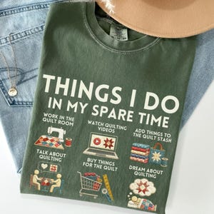 May include: Olive green t-shirt with the text "THINGS I DO IN MY SPARE TIME". The shirt features illustrations of quilting-related activities, including sewing, watching videos, and buying supplies. The shirt is displayed on a light blue denim background.