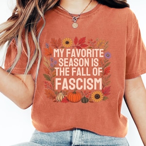 May include: A rust-colored short-sleeve t-shirt featuring a graphic print of autumn leaves, sunflowers, and pumpkins surrounding the text MY FAVORITE SEASON IS THE FALL OF FASCISM in a light cream color, centered on the front of the garment.