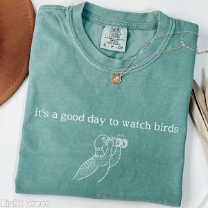 May include: Light green t-shirt with the text "it's a good day to watch birds" and a bird illustration. A silver necklace with a small pendant rests on the shirt. The shirt is labeled "Comfort Colors".