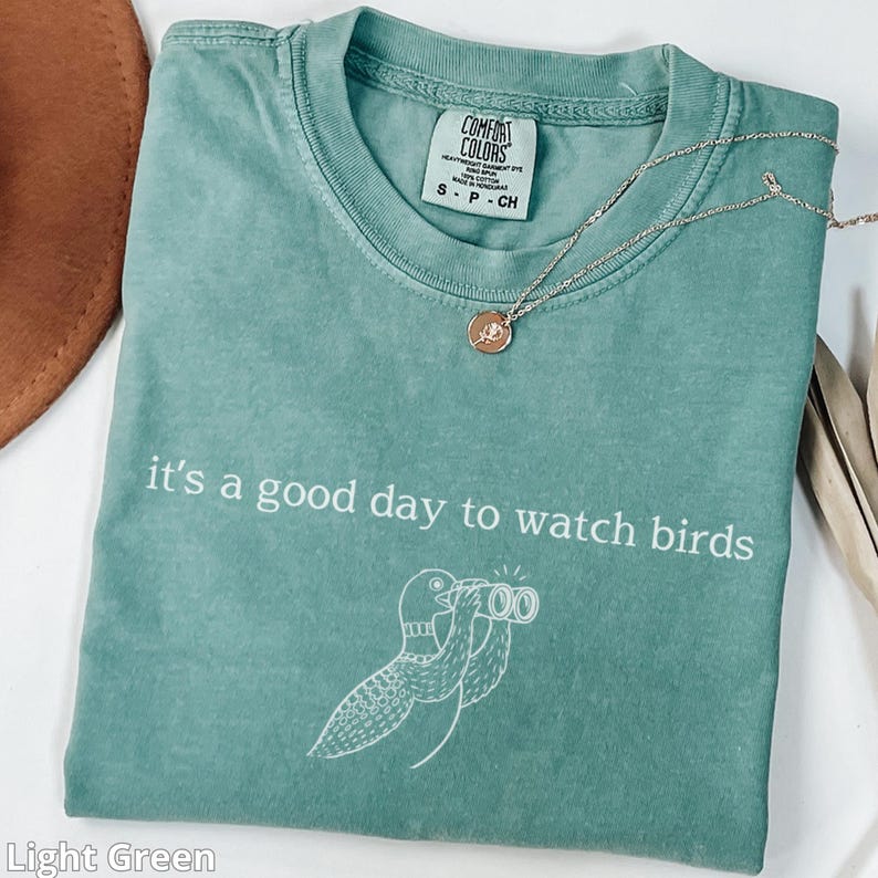 May include: Light green t-shirt with the text "it's a good day to watch birds" and a bird illustration. A silver necklace with a small pendant rests on the shirt. The shirt is labeled "Comfort Colors".