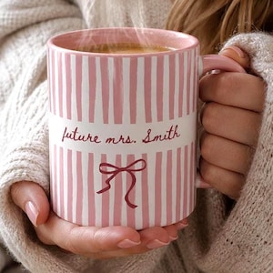 Könnte beinhalten: Eine rosa-weiß gestreifte Keramiktasse mit dem Text "future mrs. Smith" und einer Schleifen-Grafik. Die Tasse ist mit einem heißen Getränk gefüllt und hat einen rosa Griff.