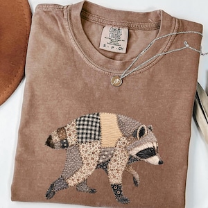 May include: A brown crew neck t-shirt featuring a graphic of a raccoon composed of various patterned fabric patches, including black and white gingham, floral prints, and star motifs. The shirt has a Comfort Colors label at the inner neckline.