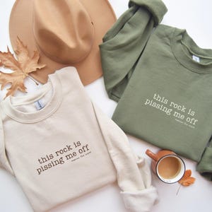 May include: Two crewneck sweatshirts, one beige and one olive green, with the phrase "this rock is pissing me off" printed on the front. A brown hat and a cup of coffee are also in the image.