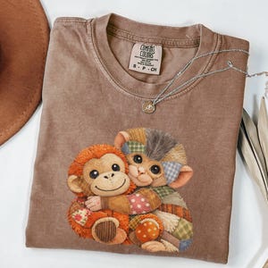 Puede incluir: Una camiseta marrón Comfort Colors con un gráfico de dos monos de patchwork abrazándose. La camiseta tiene cuello redondo y mangas cortas. Un collar plateado con un pequeño colgante está drapeado sobre la camiseta. Un sombrero marrón es visible en la esquina superior izquierda.