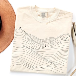 May include: A cream-coloured t-shirt featuring a black line art graphic of rolling hills, a shepherd with a staff, and a flock of sheep. The inner neck label displays the text Comfort Colors, indicating a heavyweight garment made of 100% cotton.