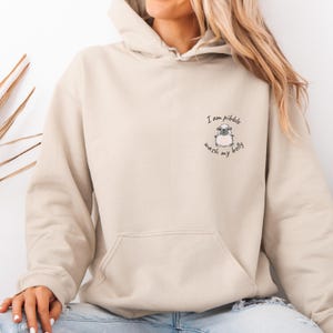 Pode incluir: Moletom bege com bolso frontal e capuz com cordão. O moletom apresenta um pequeno gráfico de um pug com o texto "I am pibble wash my belly" em escrita preta.