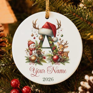 May include: A round white ceramic Christmas ornament featuring a large letter A decorated with a Santa hat, a snowman, a reindeer, pine branches, and ornaments. The text Your Name and the year 2026 are printed in red and dark green below the letter.