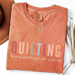 May include: A coral-colored t-shirt with the word "QUILTING" in colorful block letters. Below the word is the phrase "because stabbing people is wrong" in a cursive font. A gold necklace with a pendant is draped on the shirt.