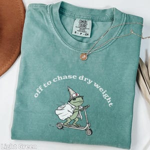 Puede incluir: Camiseta Comfort Colors verde claro con la frase "off to chase dry weight" y una rana de dibujos animados en un scooter. La rana lleva un gorro y capa de enfermera. También se ve un collar plateado con un colgante.