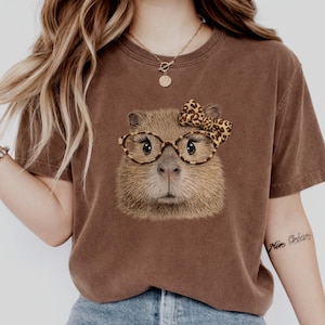 May include: Brown t-shirt featuring a capybara wearing glasses and a bow. The capybara has a detailed, realistic design with a cute expression. The glasses and bow have a leopard print pattern. The shirt is a solid brown color.