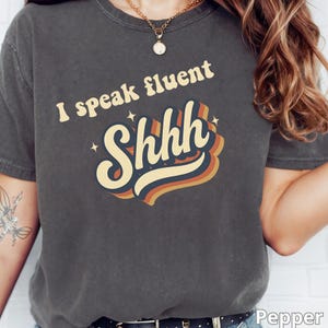Pode incluir: T-shirt cinza escuro com o texto "I speak fluent Shhh" em um design retrô. A palavra "Shhh" está em uma fonte em camadas e colorida com detalhes em laranja, amarelo e azul. A camiseta é de estilo casual.