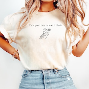 May include: Cream-colored t-shirt with the phrase "it's a good day to watch birds" printed in black. A black and white illustration of a bird looking through binoculars is below the text. The shirt is made of a soft material.