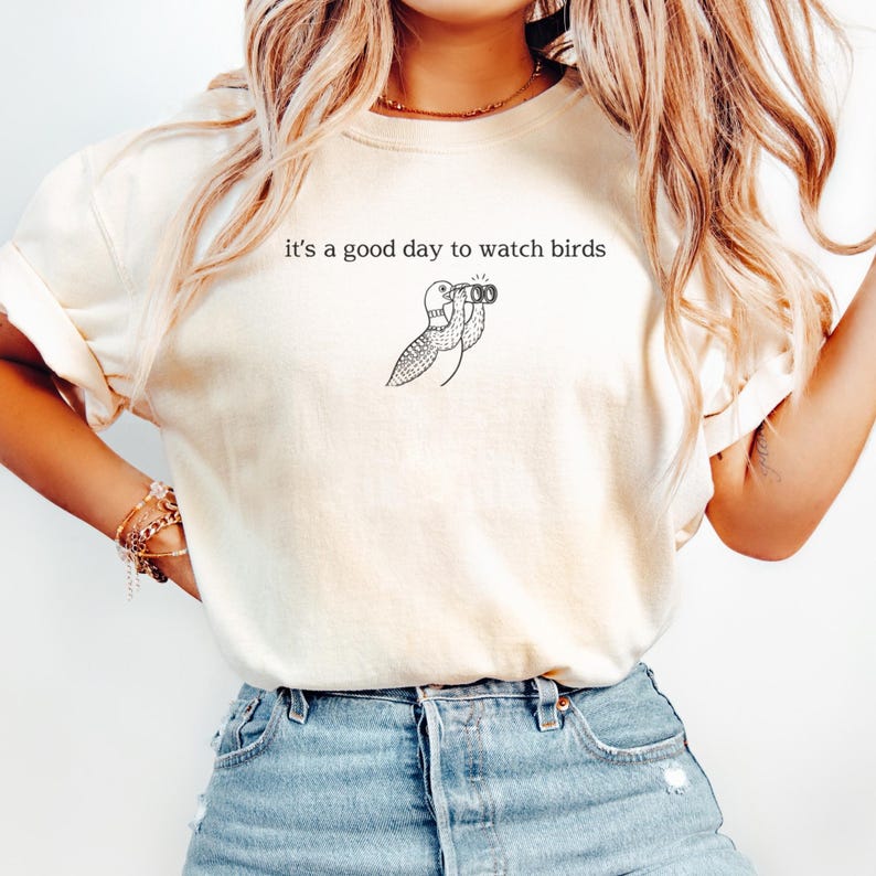May include: Cream-colored t-shirt with the phrase "it's a good day to watch birds" printed in black. A black and white illustration of a bird looking through binoculars is below the text. The shirt is made of a soft material.