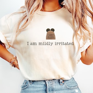 以下が含まれることがあります： 「I am mildly irritated」の黒い文字が入ったクリーム色のスウェットシャツ。黒い耳の顔の漫画の画像が特徴です。モデルはブルージーンズを着用しています。