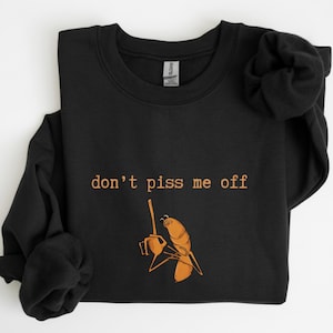 May include: Black sweatshirt with the text "don't piss me off" in a light brown color. Below the text is a light brown illustration of a shrimp holding a knife.