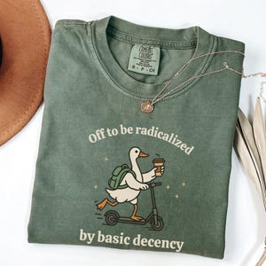 Könnte beinhalten: Grünes Comfort Colors T-Shirt mit dem Schriftzug "Off to be radicalized by basic decency" und einer Grafik einer Ente auf einem Roller, die Kaffee hält. Eine goldene Halskette und ein brauner Hut sind ebenfalls zu sehen.