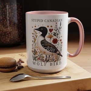 May include: White ceramic mug with a pink interior and handle. The mug features a detailed illustration of a loon bird surrounded by flowers, with the text "STUPID CANADIAN WOLF BIRD" printed on the front.