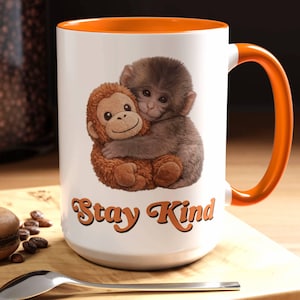 May include: White ceramic mug with an orange interior and handle. The mug features an illustration of two monkeys hugging, with the text "Stay Kind" in orange script. The mug is on a wooden surface.