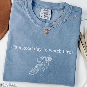 May include: Blue jean colored t-shirt with the text "it's a good day to watch birds" and a bird graphic. A gold necklace with a pendant is on the shirt. The shirt is labeled Comfort Colors, size S-P-CH.