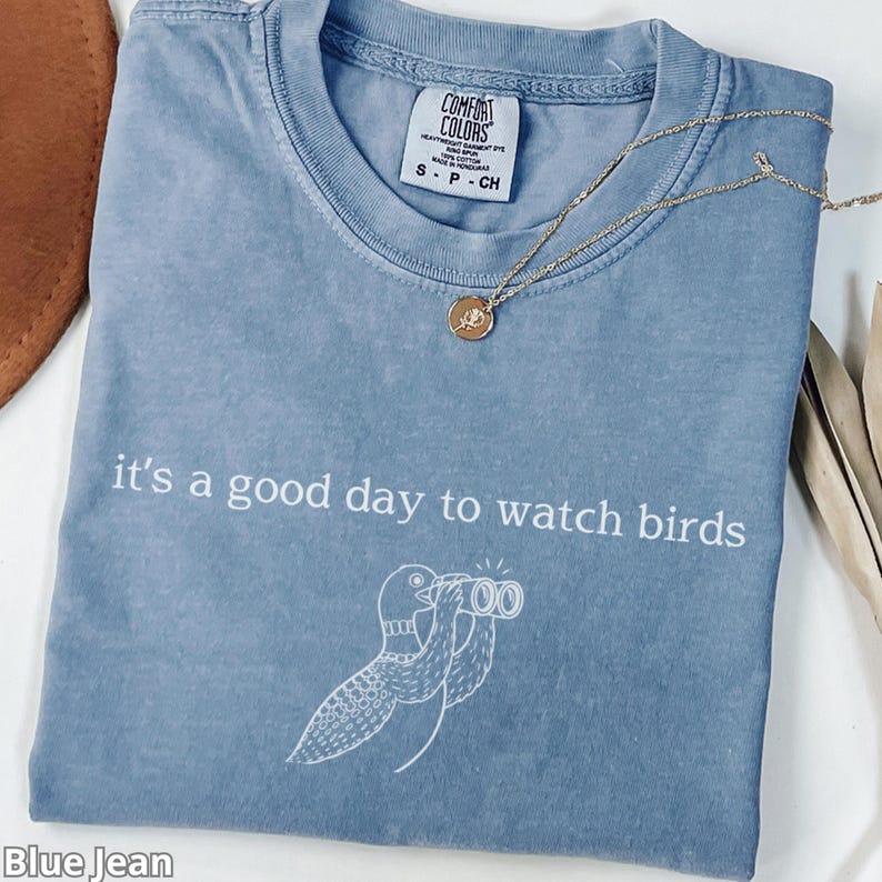 May include: Blue jean colored t-shirt with the text "it's a good day to watch birds" and a bird graphic. A gold necklace with a pendant is on the shirt. The shirt is labeled Comfort Colors, size S-P-CH.