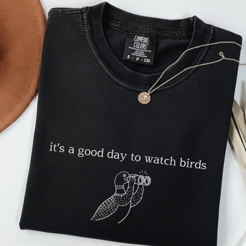 May include: Black t-shirt with the phrase "it's a good day to watch birds" and a bird illustration. The shirt has a crew neck and short sleeves. A gold necklace with a pendant is on the shirt.
