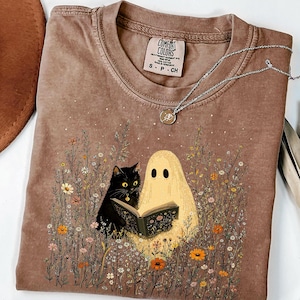 Puede incluir: Una camiseta marrón de manga corta con un gráfico de un gato negro y un fantasma beige leyendo un libro juntos en un campo de flores silvestres. La camiseta tiene cuello redondo y una etiqueta de la marca Comfort Colors en el interior.