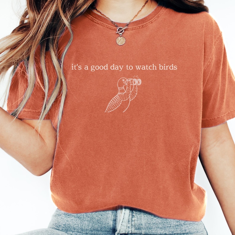 May include: Burnt orange t-shirt with the phrase "it's a good day to watch birds" and a line drawing of a bird with binoculars. The shirt is a comfortable, casual style.