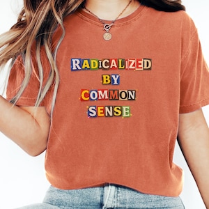Op de afbeelding: Verbrand oranje t-shirt met de tekst "RADICALIZED BY COMMON SENSE" in kleurrijke blokletters. Het shirt heeft een ronde hals. De tekst is in drie regels gerangschikt.