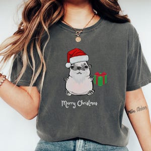 Pode incluir: T-shirt cinzenta escura com um desenho de pug a usar um gorro e barba de Pai Natal, segurando uma caixa de presente verde. As palavras "Merry Christmas" estão impressas abaixo do pug. A t-shirt é de estilo casual.