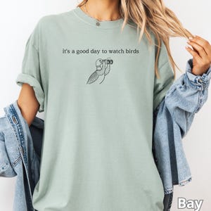 May include: A light green t-shirt with the phrase "it's a good day to watch birds" and a black illustration of a bird using binoculars. The shirt is a solid color and has a relaxed fit.