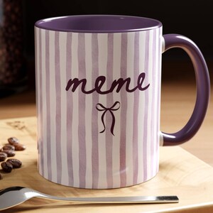 May include: A ceramic mug with a purple interior and handle. The exterior features vertical stripes in white and lavender, with the word "meme" and a bow graphic in dark purple.