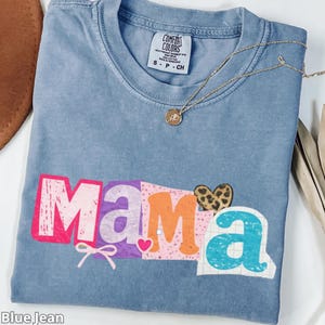 May include: Blue jean colored t-shirt with the word "Mama" in colorful block letters. The shirt has a small bow and heart graphic. A gold necklace with a pendant is draped across the shirt.