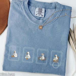May include: Blue jean Comfort Colors t-shirt with four postage stamp-style illustrations of a goose playing a drum. A gold necklace with a pendant rests on the shirt. The shirt is labelled S-P-CH.