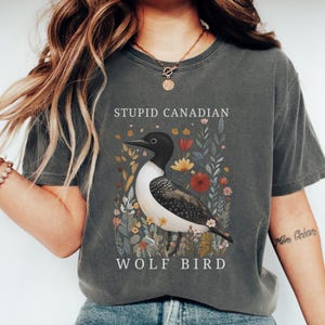 May include: Dark grey t-shirt featuring a detailed illustration of a loon bird surrounded by flowers. The text "STUPID CANADIAN" is above the bird, and "WOLF BIRD" is below. The shirt has short sleeves and a crew neck.