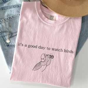 May include: A light pink t-shirt with the phrase "it's a good day to watch birds" and a cartoon bird with binoculars. The shirt is folded and placed on top of blue jeans and a necklace.