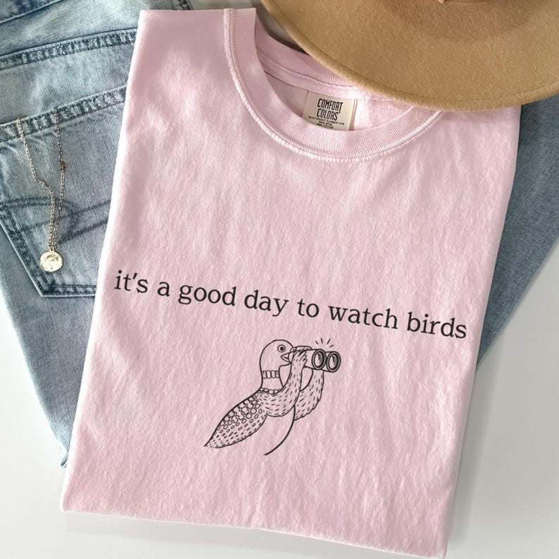 May include: A light pink t-shirt with the phrase "it's a good day to watch birds" and a cartoon bird with binoculars. The shirt is folded and placed on top of blue jeans and a necklace.