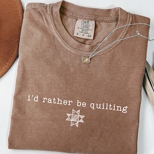 May include: A brown short-sleeved t-shirt with a crew neckline and a Comfort Colors label. The shirt features the white text "i'd rather be quilting" above a white geometric star graphic. A thin silver chain necklace rests on the fabric.