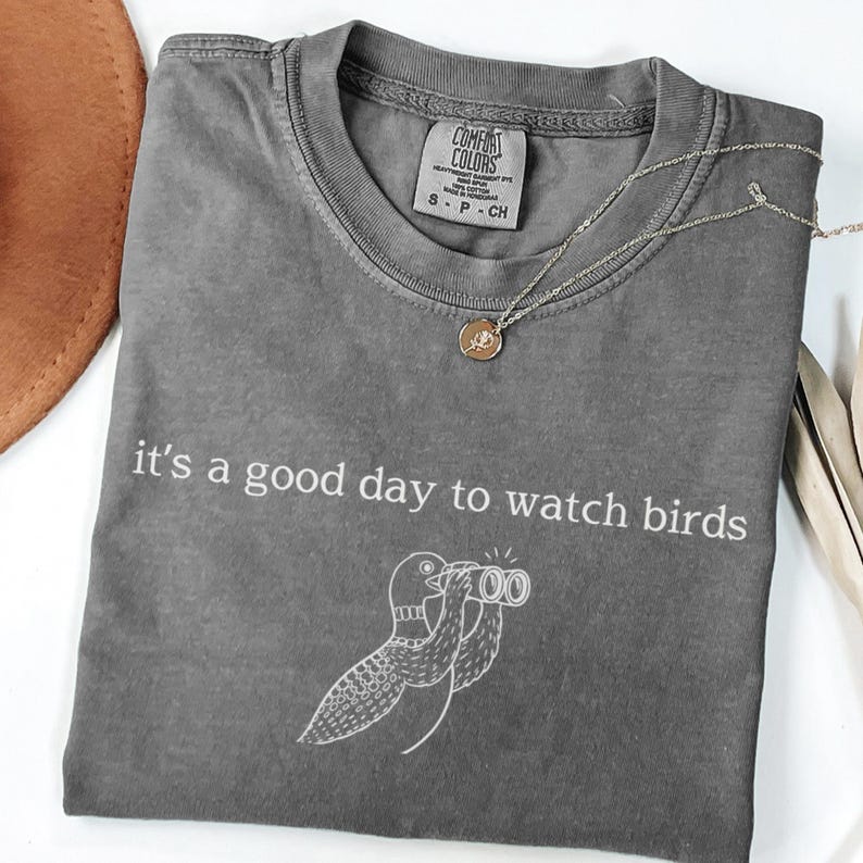 May include: A gray Comfort Colors t-shirt with the text "it's a good day to watch birds" and a bird illustration. A gold necklace with a pendant is on the shirt. The shirt is folded.