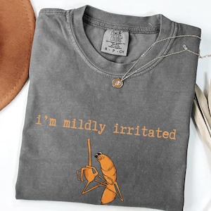 Puede incluir: Camiseta gris Comfort Colors con la frase "I'm mildly irritated" en letras naranjas. Debajo del texto hay un dibujo animado de un camarón naranja con el dedo corazón levantado. Un collar dorado con un pequeño colgante está sobre la camiseta.