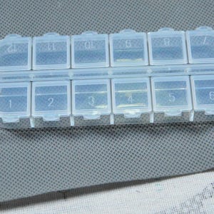 May include: A clear plastic pill organizer with twelve numbered compartments. The rectangular case has a hinged lid for each compartment, designed for daily or weekly medication management. The compartments are labeled with numbers 1 through 12.