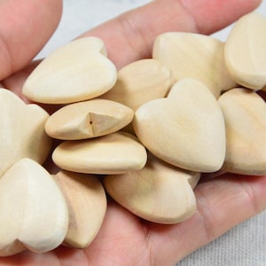 May include: A handful of light-colored wooden heart-shaped objects. The hearts are smooth and appear to be unfinished, with a natural wood grain. The objects are held in a person's hand.