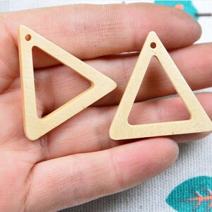 Puede incluir: Dos dijes triangulares de madera de color claro con un acabado liso. Cada pieza presenta un centro hueco y un pequeño orificio circular en la punta superior, diseñado para su fijación. La veta de la madera natural es visible en las superficies.