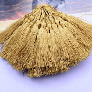May include: A collection of golden tassels, each with a looped top and long, flowing strands. The tassels are arranged in a fan shape, creating a textured and decorative element. The background is a light purple surface.