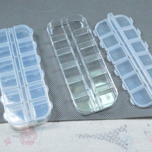 Peut inclure: Trois piluliers en plastique transparent. Chaque organisateur comporte plusieurs petits compartiments pour ranger des pilules ou de petits objets. Les organisateurs sont rectangulaires avec des bords arrondis.