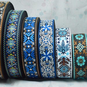 Könnte beinhalten: Eine Sammlung von sieben Rollen gewebtem Jacquardband, die in einer Reihe angeordnet sind und komplizierte florale und geometrische Stickmuster in verschiedenen Farbkombinationen aus schwarzem, blauem, weißem, orangefarbenem und türkisfarbenem Garn aufweisen.