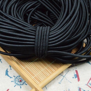 May include: A coiled length of black elastic cord featuring a smooth, rounded texture. The cord is wound into a dense, circular bundle and secured with a central wrap of the same black material, creating a uniform and organized appearance.