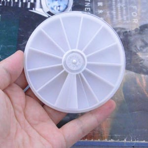 May include: A white, circular container with multiple compartments, possibly for storing small craft items. The container is held in a hand and has a central hub with radiating segments. The plastic is opaque.