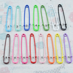 May include: A collection of fifteen safety pins in various colors, including blue, green, white, black, brown, pink, red, orange, yellow, and purple. Each pin is numbered from 1 to 15.