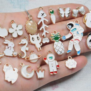 May include: A collection of enamel charms in various shapes, including a sheep, flower, rabbit, seahorse, cat, astronaut, pineapple, panda, and cloud. The charms are primarily white with gold accents and are suitable for jewelry making or crafting.