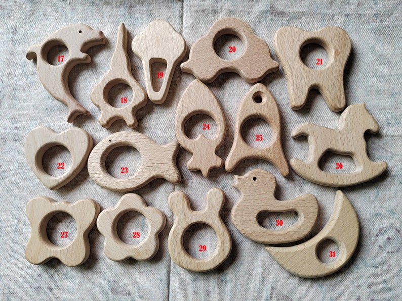 May include: A set of 14 wooden teethers in various shapes, including a dolphin, ice cream cone, car, tooth, rocket, fish, heart, rocking horse, duck, moon, flower, bunny, and star. The teethers are all made of natural wood and have a smooth, polished finish.